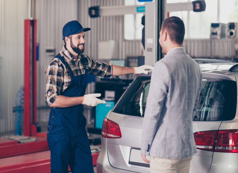 Vehicle breakdown, check, inspection, engineering concept. Cheerful specialist in blue overall, cap, checkered shirt enjoying conversation with client in formal outfit in work shop